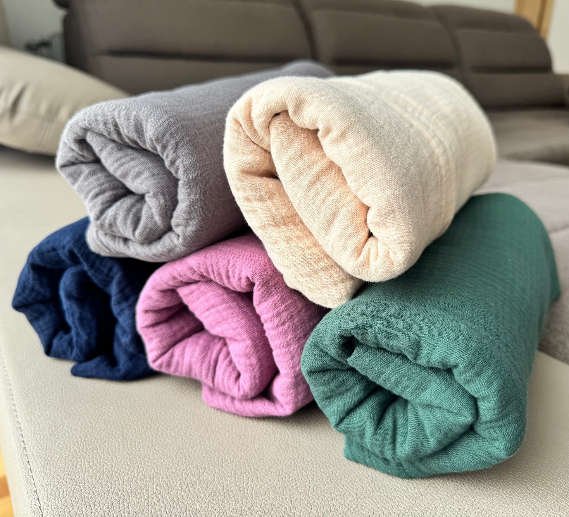 Stack of folded muslin cotton scarf wrap blankets in various colors on a sofa.