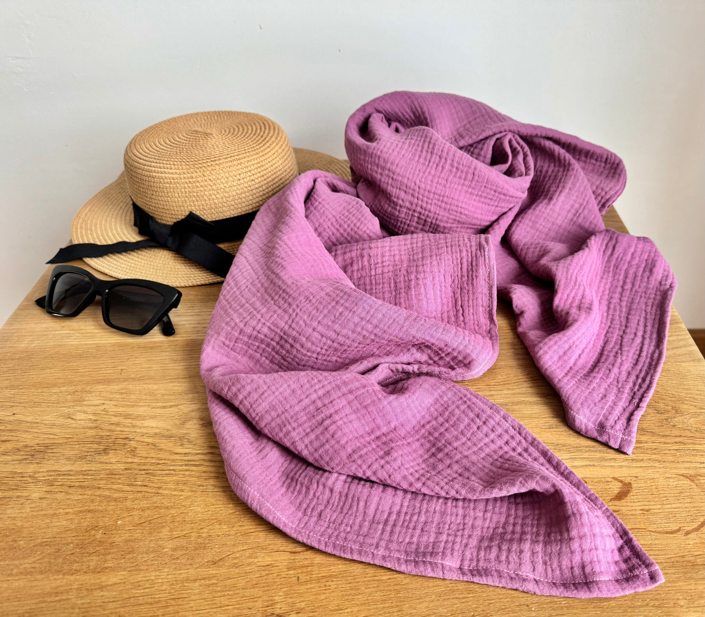 Dusty rose muslin cotton scarf wraop, straw hat, and sunglasses on a wooden surface