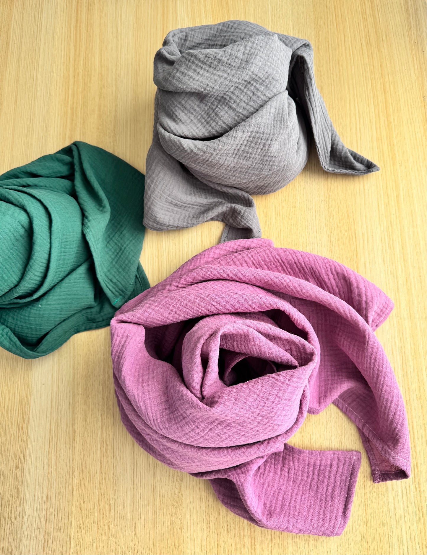 Three rolled-up scarf wraps in emerald green, graphite gray, and dusty rose pink on a wooden surface